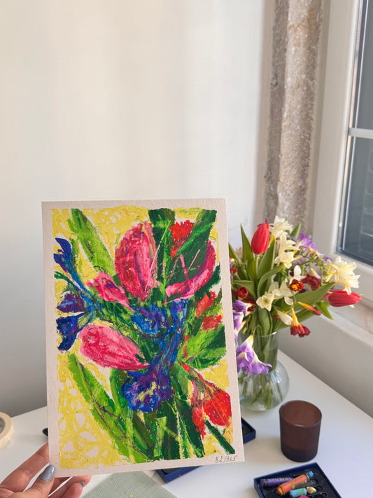 Colorful floral painting held by a person with a real flower arrangement in the background.
