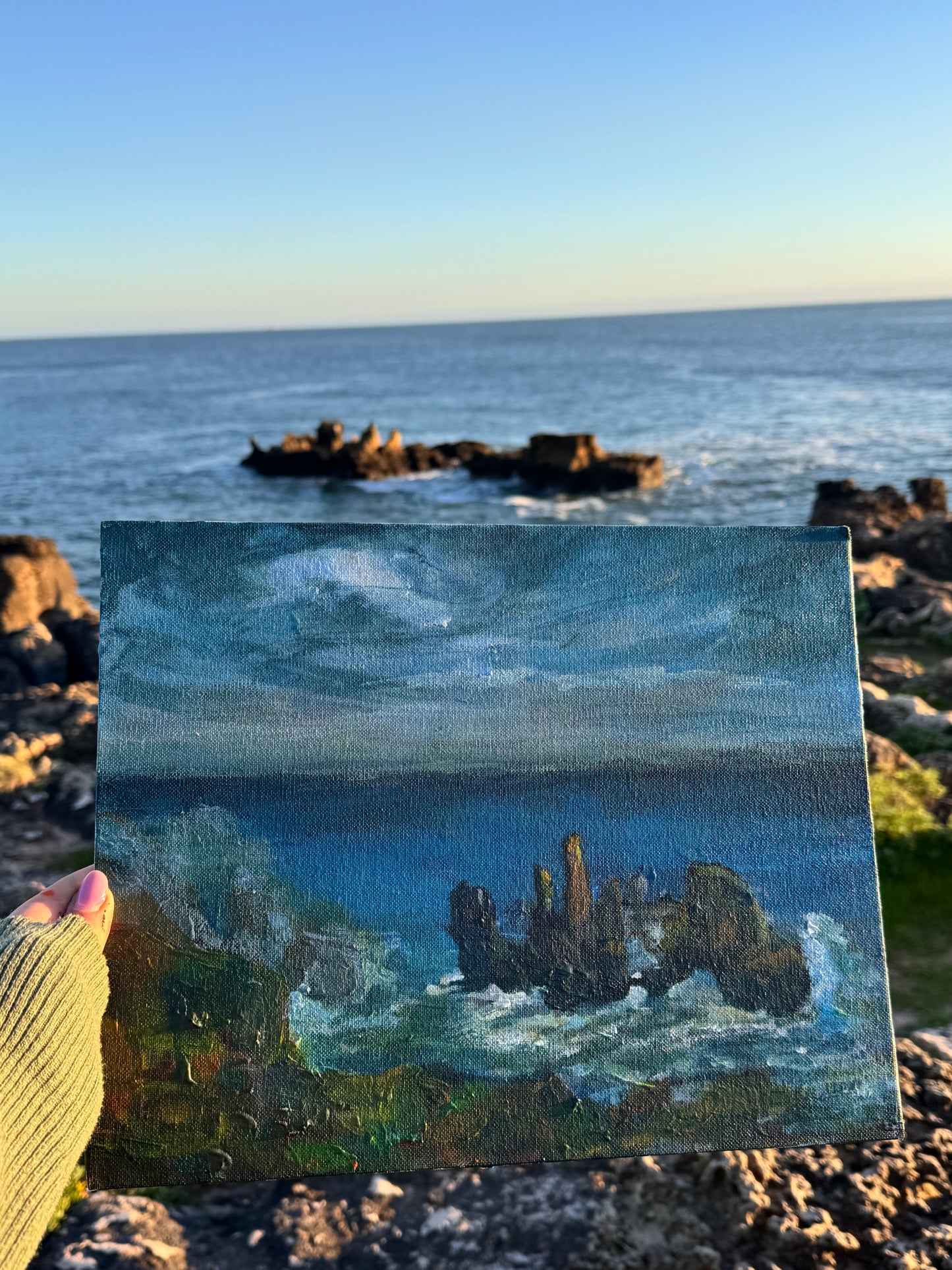 Small painting of a coastal scene held in front of a real coastal landscape.