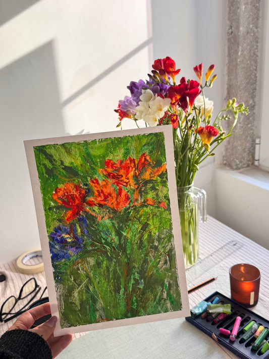 Person holding a framed painting of flowers next to a vase with real flowers on a table.
