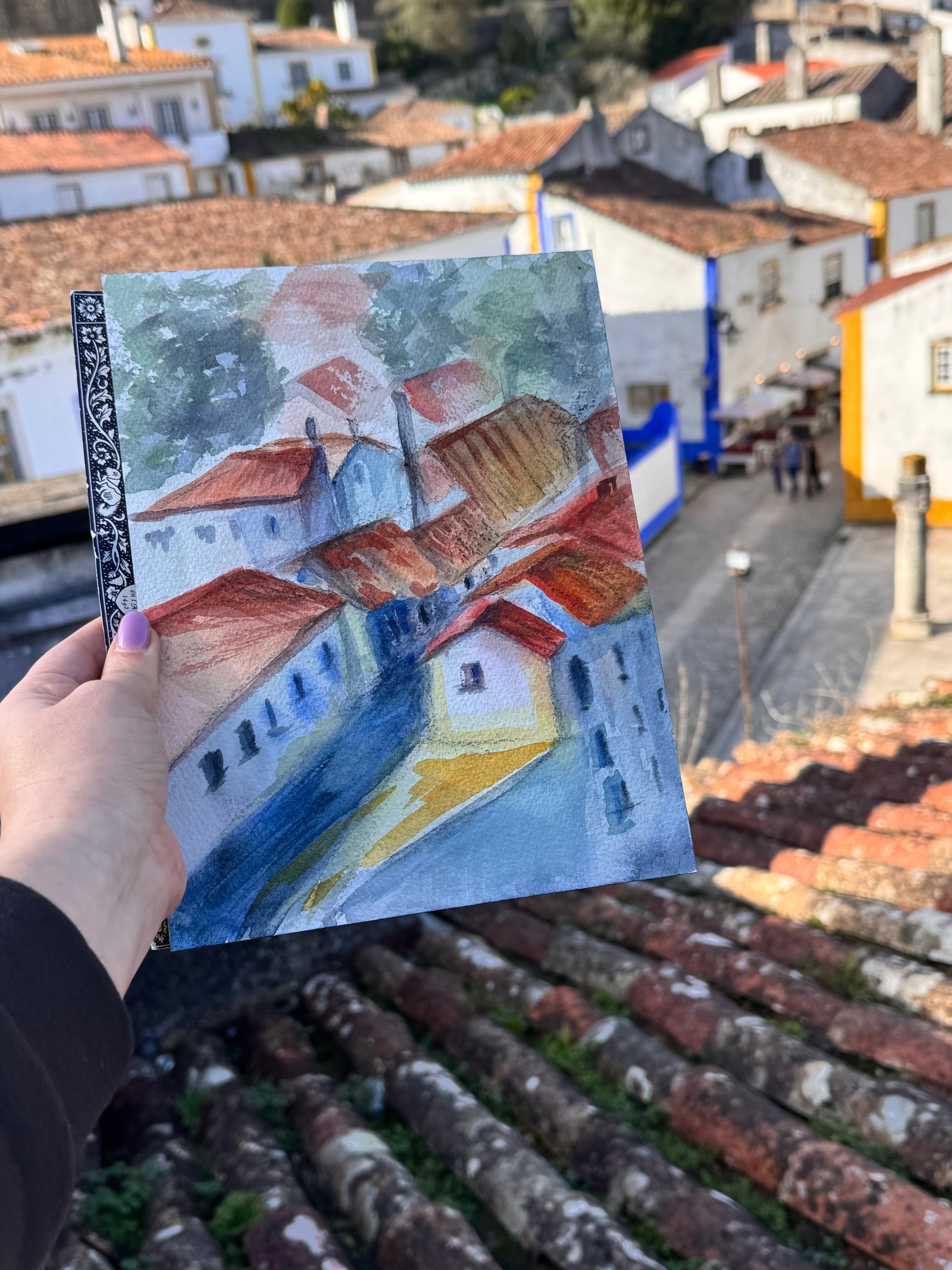 a small painting of a village scene with rooftops, with a real-life village in the background.